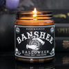 a Lit witchy scented candle with a black label with the graphic of witchy hands holding a crystal ball on it and says "Banshee" 