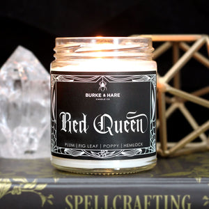 A lit candle set in a witchy scene with spell books and a crystal. Black  candle label has a spiderweb border and says Red Queen in gothic font