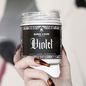witchy hand with long fingernails holding a sandalwood scented candle with a black label and spiderweb border. Candle reads "violet" in gothic font