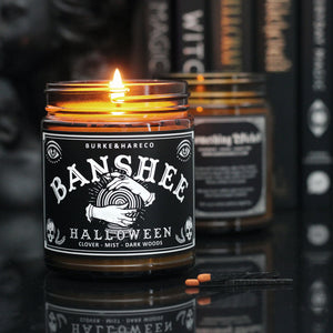 a Lit witchy scented candle with a black label with the graphic of witchy hands holding a crystal ball on it and says "Banshee" 