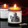 A  lit candle in an amber jar sits in a dark room. The candle's label features the image of a handsome pale vampire and says "Dracula" in gothic font and scent notes calla lily, red rose, earth are listed in metallic gold letters.