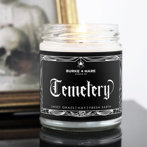 a lit cemetery scented candle infront of a vintage photo and gothic home decor scene. Candle has a gothic label that has white spiderweb border and white text that reads "Cemetery"