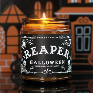 lit  Halloween scented candle that has a black label with graphic skeletons on it and says "Reaper" in white ouija lettering