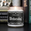 Gothic scented candle with black label and spiderweb border that reads Wednesday in gothic font. The candle is lit and on a shelf in front of witchy books.