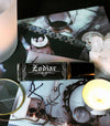 zodiac perfume oil with a black label and white text on a witchy background of gothic photos