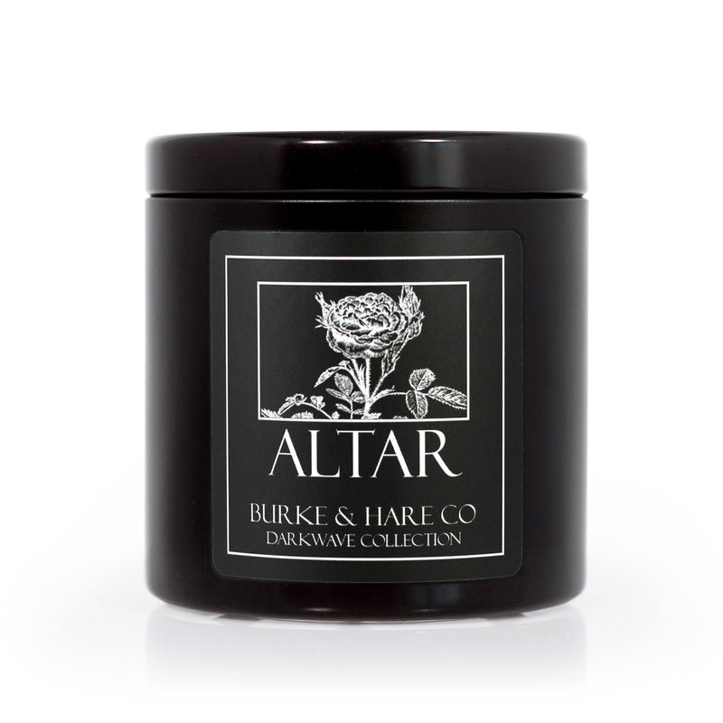 dark home interior with a lit Vampire inspired scented candle in a black tin with a black label featuring an illustrated rose and white text that says "Altar"