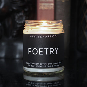a lit candle that smells like books in a dark academia scene with books in the background. Candle has a black label with white text that says Poetry. 