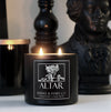 dark home interior with a lit Vampire inspired scented candle in a black tin with a black label featuring an illustrated rose and white text that says "Altar"