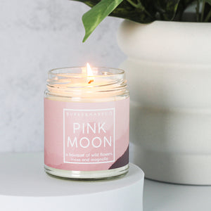 a lit floral scented candle on a kitchen counter in front of a houseplant. Candle has a pastel pink label and white text that says Pink Moon